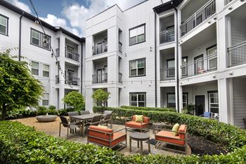 A patio with a table and chairs is surrounded by a well-kept garden in front of a white apartment building.